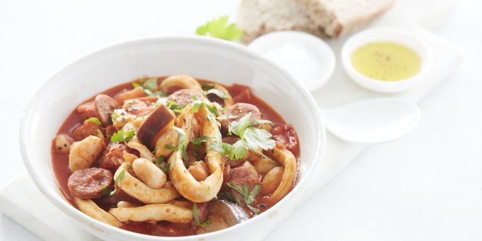 Een goed gevuld wit bord met het stoofpotje staat klaar om aan te vallen. De kleur is rood van de vele tomaten en chorizo die zijn gebruikt. Het bordje staat op een marmere snijplank waar ook brood en een klein potje olijfolie op staat. 