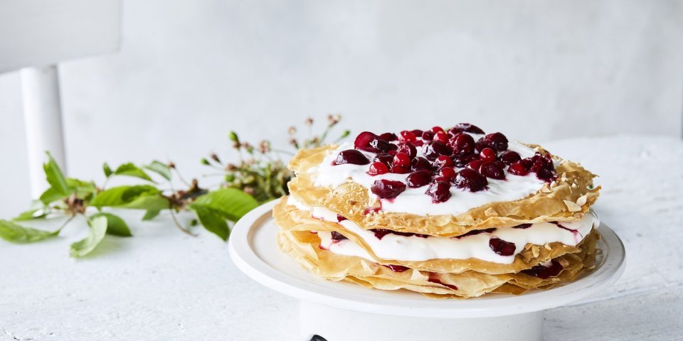 Grote ronde millefeuille met kersen en verse roomkaas. Deze staat op een witte taartschaal op een bleke houten tafel. Op tafel ligt ook een takje groen voor extra sfeer. 