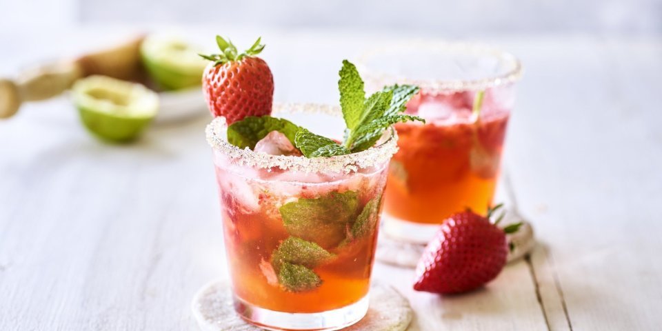 Twee roodgekleurde mojito's op de tafel, afgewerkt met verse munt en nog een aardbei. Alleen al kijken is een feestje, tijd om die cocktail te benuttigen. Het glad kreeg zelfs een suikerrandje. 