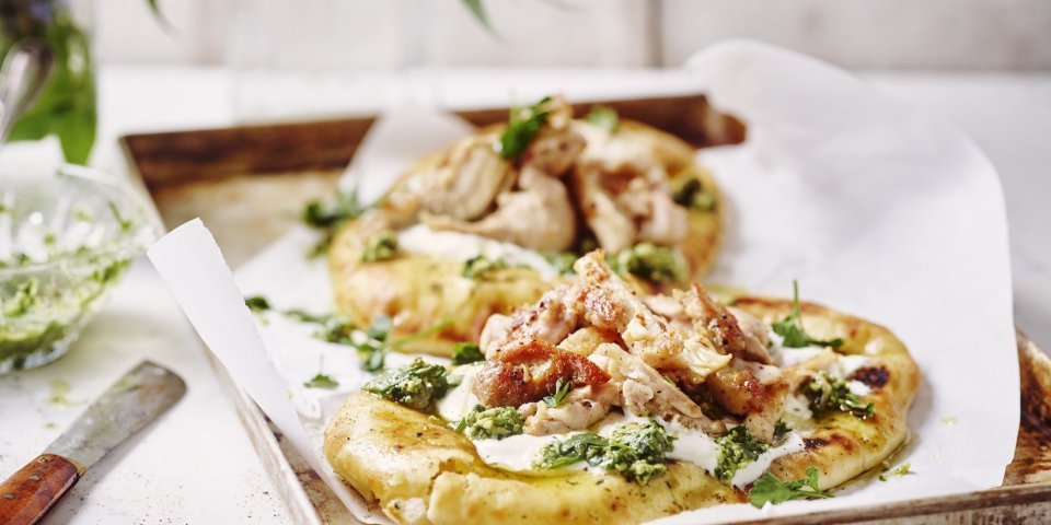 Twee naanbroodjes liggen op een metalen ovenplaat. Ze zijn belegd met yoghurt, stukjes gebakken kip en afgewerkt met salsa verde. De ovenplaat staat op een witte houten tafel.