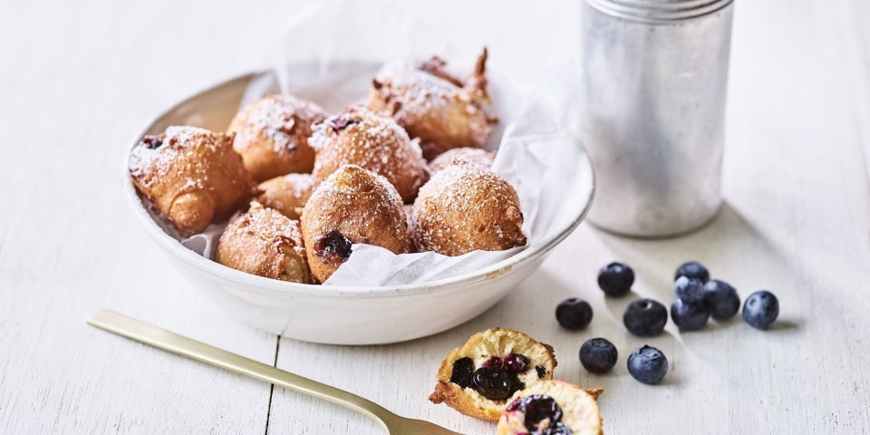 In een diep wit bord ligt een wit boterpapiertje met hierin enkele oliebollen. Er is eentje uitgeprikt en ligt op tafel doormidden gesneden, hier zie je duidelijk de blauwe bessen in zitten. Er is bloemsuiker over de oliebollen gestrooid voor een extra zoete toets. 