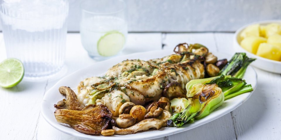 Ovengebakken zeeduivel ligt op een groot wit bord, geserveerd met gegrilde paksoi en champignons. Het gerechtje wordt geserveerd met gekookte aardappelen. Bovenaan staat ook ijskoud water met limoenschijfjes. 