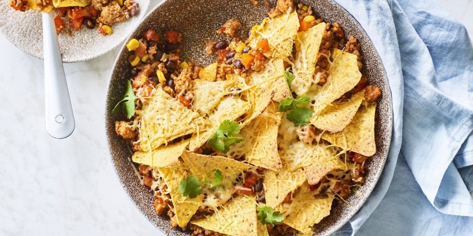 Onderaan de ronde ovenschaal ligt de chili con carne met bovenop de nachos. Er is al een portie uitgeschept op een wit bordje en staat langs de ovenschaal. Er ligt ook een blauwe servette en enkele blaadjes kroiander voor de kleur.