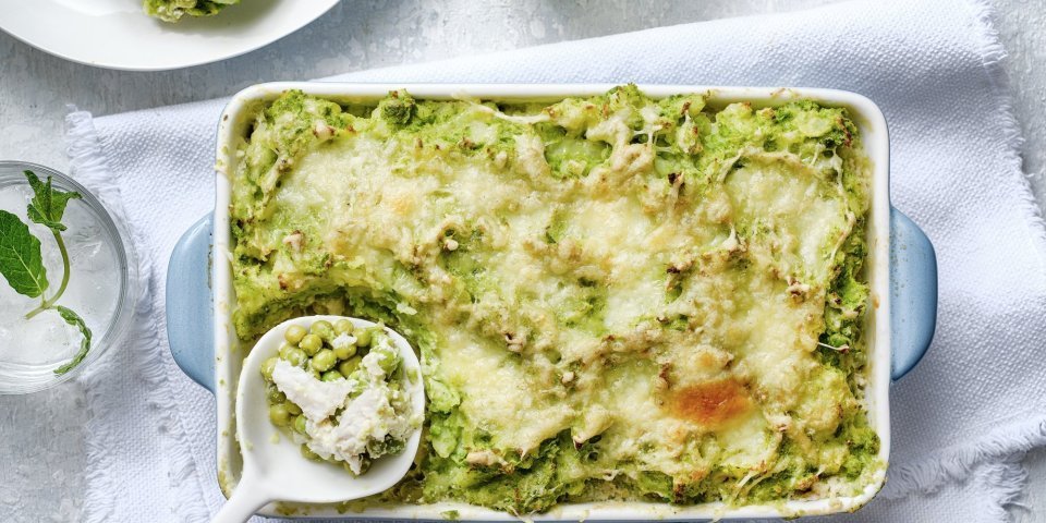 Eten is klaar! De ovenschotel staat op een witte dikke doek en een porseleine lepel schept de eerste portie uit de schotel. De parmentier heeft een mooie frisse groene kleur door de vele erwtjes. 