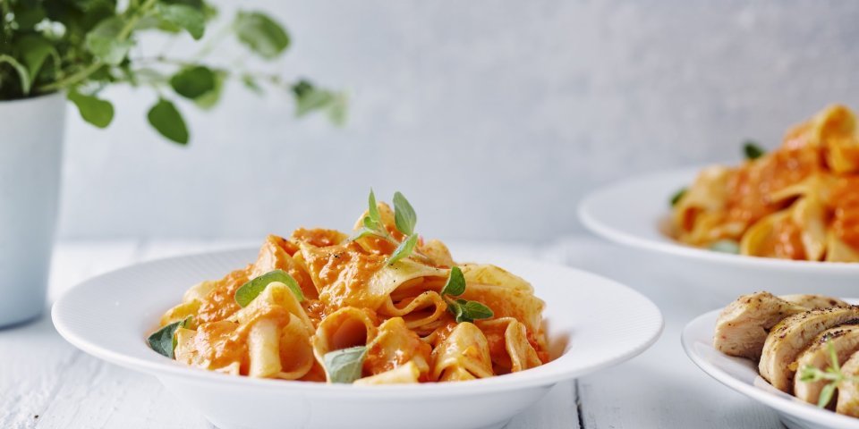 Een portie pasta met homemade paprikasaus staat klaar op een witte houten tafel. Een vers plantje oregano staat achteraan in beeld. 