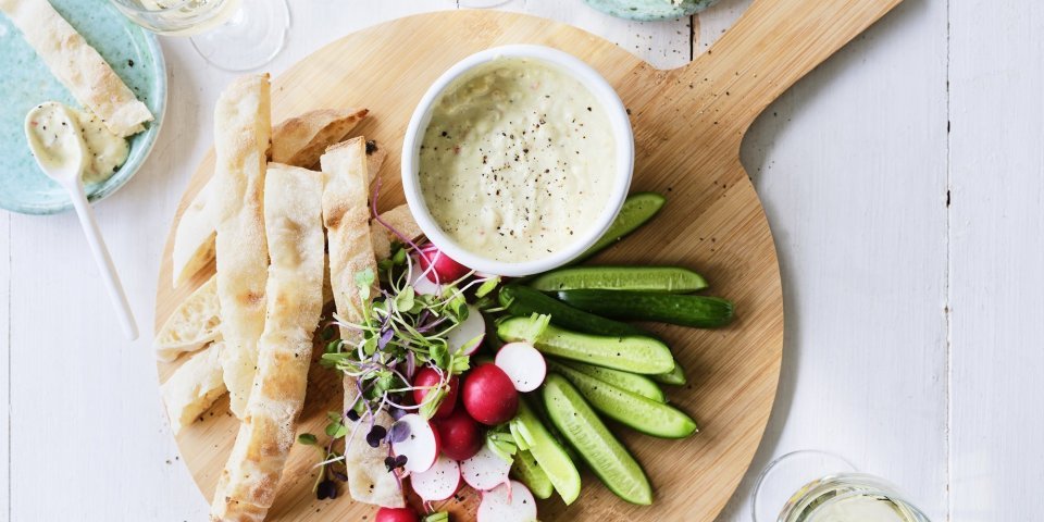 Op tafel staat een ronde houten snijplank met hierop een wit potje gevuld met onze pikante dip van yoghurt en witte bonen. Je kan de yoghurtdip gebruiken met stukjes snackkomkommer, brood en radijsjes in ons geval. Rond de plank staan enkele glazen witte wijn en helderblauwe broodbordjes. 