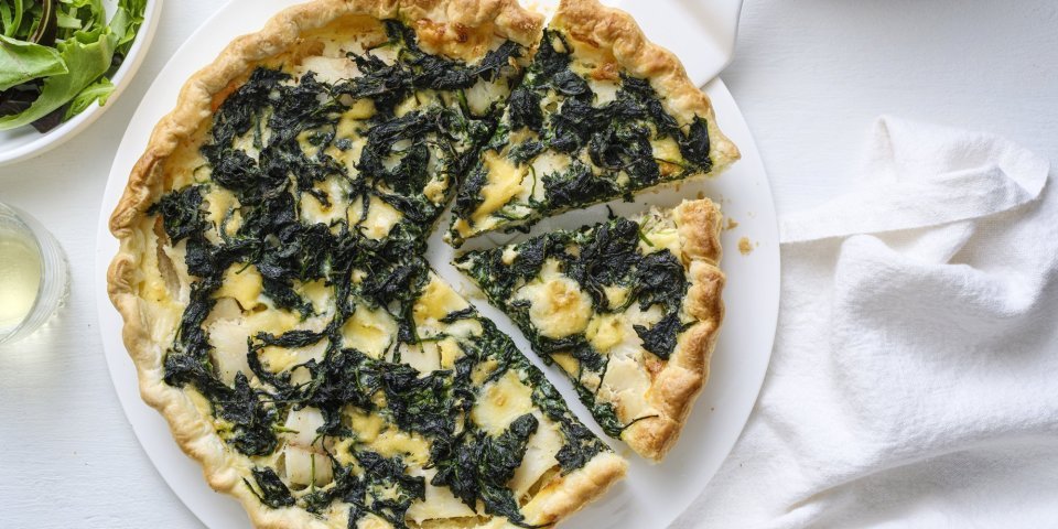 Op een taartschaal ligt de aangesneden quiche. Er is een witte taartschep onder één stukje quiche om naar een bordje te gaan. Langs de quiche staat ook nog een portie gemengde sla, voor nog een extra portie groenten. 