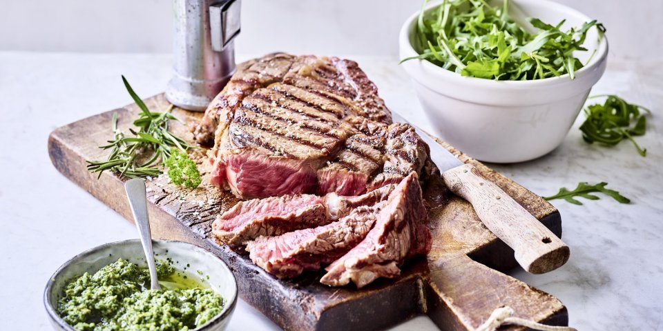 Een grote gegrilde steak ligt op een houten snijplank aangesneden. Er staat een potje homemade rucolapesto langs om meteen bij te scheppen bij je reepje vlees. Er staat ook een metalen pepermolen langs wie het graag wat pittiger wil.