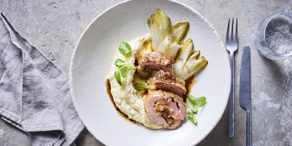 Een stukje rollade van varkensvlees ligt in een wit groot bordje op een smeuige pastinaakpuree met gebakken witloofstronkjes. 