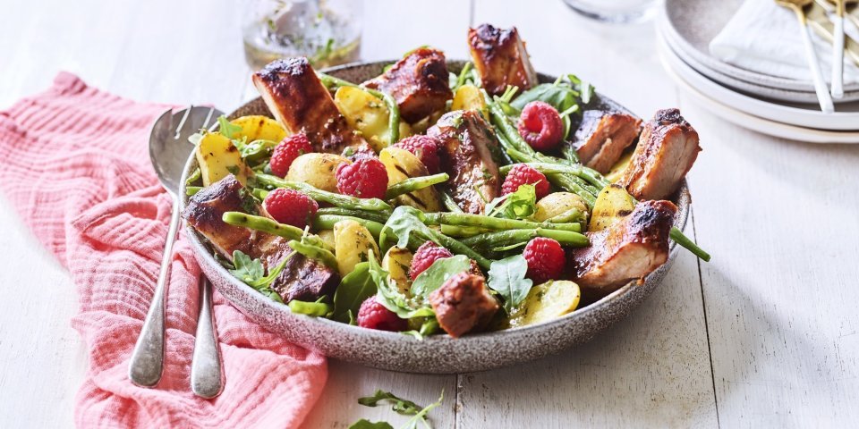 Een grote serveerschaal is gevuld met de aardappelsalade. Je ziet de framboosjes, boontjes en ribbetjes in de salade zitten. Er staat een torentje bordjes klaar met bestek om de salade te verdelen en op te eten. 