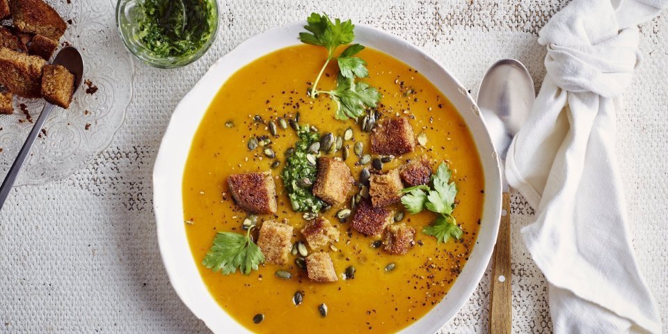 Op tafel staat een wit soepbord gevuld met heerlijk oranje pompoensoep. De soep is afgewerkt met verse groene kruiden, pompoenpitten en croutons van zuurdesembrood. Er ligt een witte linnen servette langs, geknoopt, en een soeplepel. 