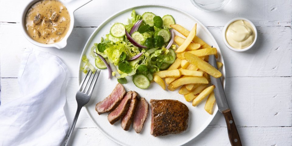 Een echte Vlaamse klassieker die smaakt voor jong & oud, een steak mooi gebakken met heerlijke goude frietjes en een salade. De champignonsaus staat langs het bord in een sauspotje. Bestek en servette liggen langs het bord klaar om te starten. 