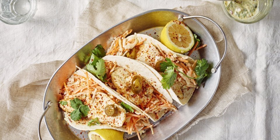 Drie wraps liggen gepresenteerd in een ovenschotel van inox. Deze staat op een gedekte tafel met wit linnen kleed. Er liggen twee citroenhelften bij de tortilla's en verse koriander. 