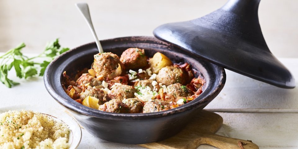 Zwartkleurige tajine met gehaktballetjes van lamsvlees, afgewerkt met verbrokkelde feta. Er staat een portie couscous naast de tajine. 