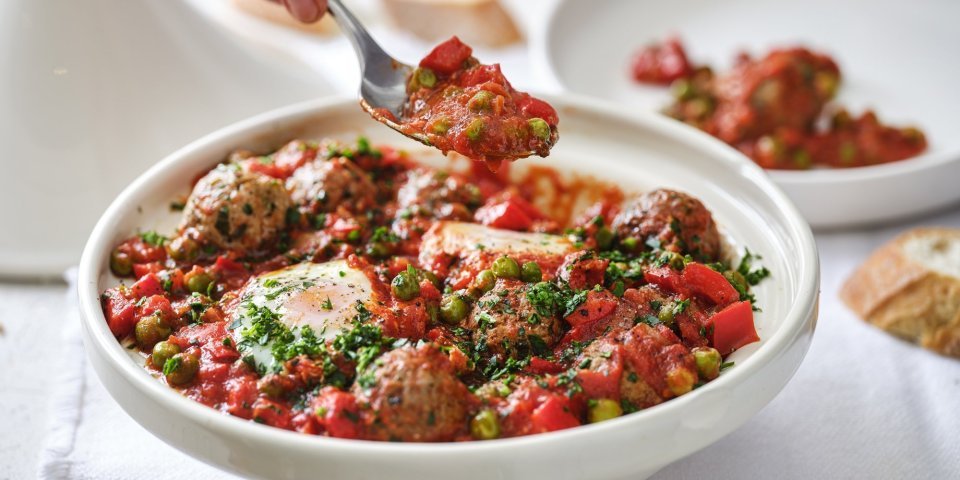 Op tafel staat een witte tajine heerlijk gevuld met gehaktballen in een rijke tomatensaus met eitjes. Er wordt net een portie uitgeschept. 