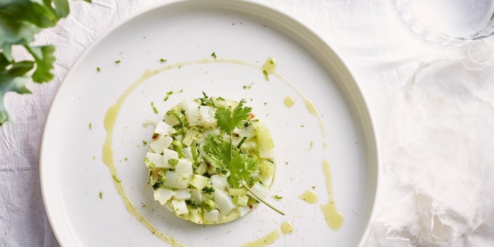 Op tafel ligt een mooi wit linnen tafelkleed, waarop het witte bord staat. Hierop ligt de tartaar van zeekat met komkommerstukjes, lente-ui en koriander. We voegden ook wat chili toe voor extra pit en als afwerking nog wat olijfolie en een korianderblaadje. 