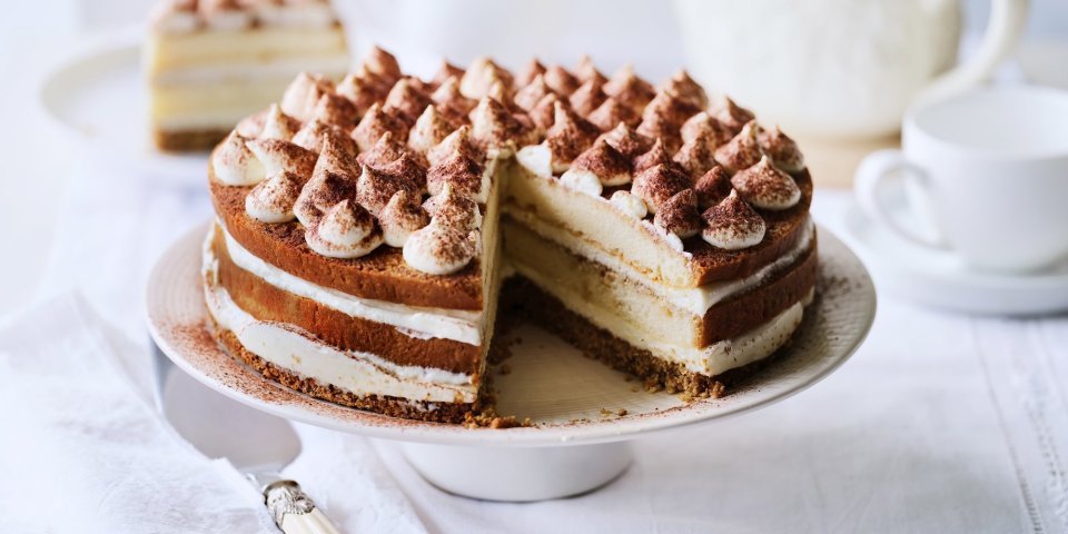 Op tafel staat een prachtige taart, het is een twist op de klassieke tiramisu. Hij staat op een witte taartschaal, en is afgewerkt met toefjes mascarpone en cacaopoeder. In de achtergrond staat een koffiekan en tasjes koffie.