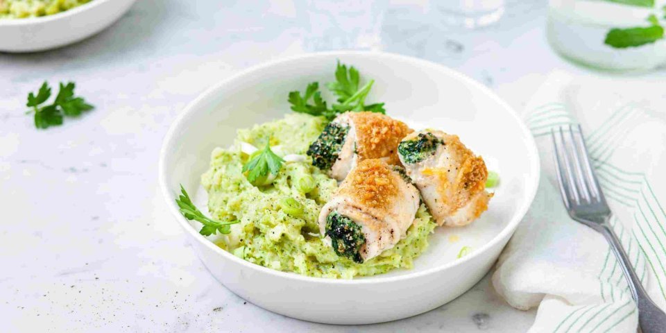 Gevulde tongrolletjes met spinazie en ricotta liggen op een groene puree van broccoli en aardappel. Ze liggen in een diep bord waarbij een karaf- en waterglas langs staat.