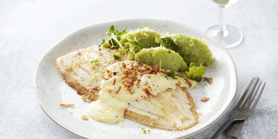 Heerlijk gebakken tongschar ligt op een wit bord met de broccoliepuree naast. Het sausje is over de vis gedresseerd met enkele gefrituurde uitjes. Bestek en een glaasje witte wijn werken het beeld af. 