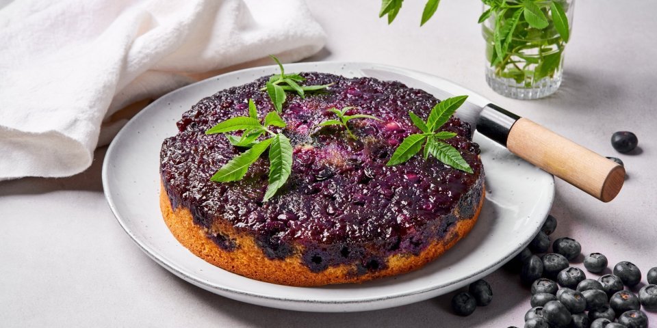 Op tafel staat een soort van tarte tatin, maar eentje met blauwe bessen en cakebeslag. Er ligt een mes langs om aan te snijden en enkele blaadjes verbena. 