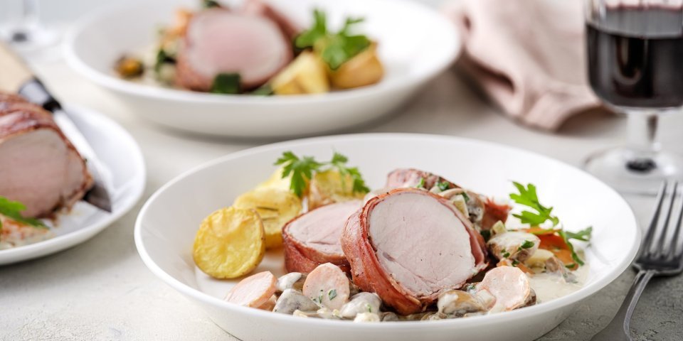 Op tafel staan twee porties uitgeschept. Het heerlijk bereide varkenshaasje ligt op de romige champignonsaus met plakjes wortel. Er liggen ook ovengeroosterde aardappeltjes bij. Langs het bord ligt een servette, glaasje rode wijn en bestek. 