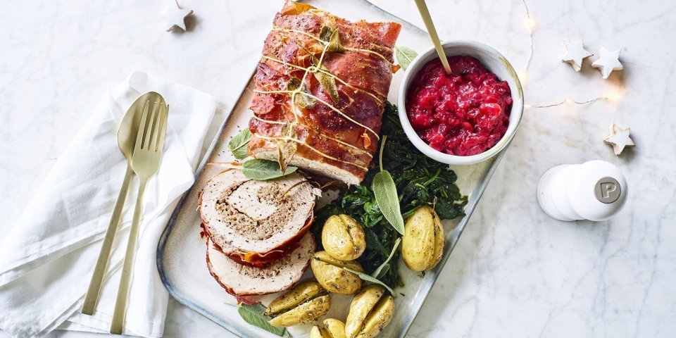 Een mooi aangesneden varkensrollade ligt op een rechthoekig dienblad. De aardappeltjes werden net niet doormidden gesneden alvorens ze de oven in gingen met een kruidentakje. De veenbessencompote staat langs de rollade die zorgt voor een intense kleur in deze feestmaaltijd. Goud bestek ligt langs het gerecht. 