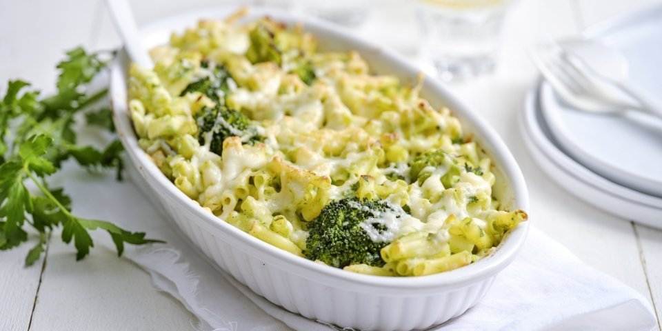 Een grote ovenschaal staat op een houte witte tafel. Hij staat op een wit lapje stof. De ovenschotel is gevuld met macaroni, broccolie en witte bloemkoolsaus. Er staan enkele bordjes langs met wit bestek om direct aan te vallen. 