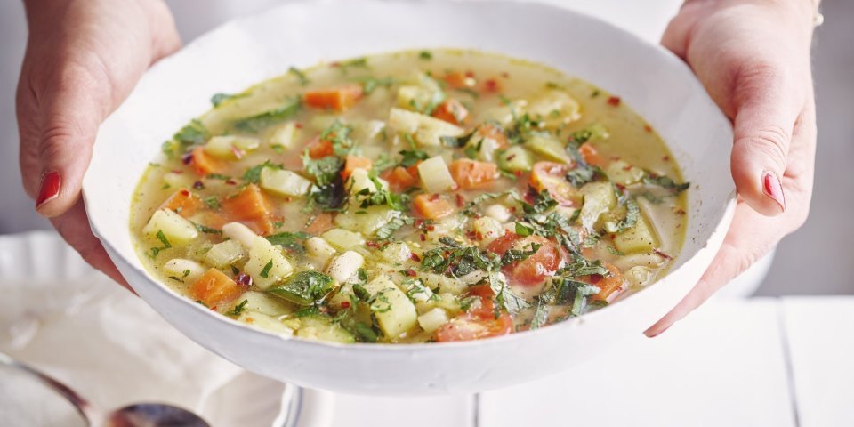 Een vegetarische minestrone met venkel, witte bonen, aardappel en verse kruiden geserveerd in een diep wit soepbord. Een vrouw met mooi rood gelakte nagels draagt het bord. 