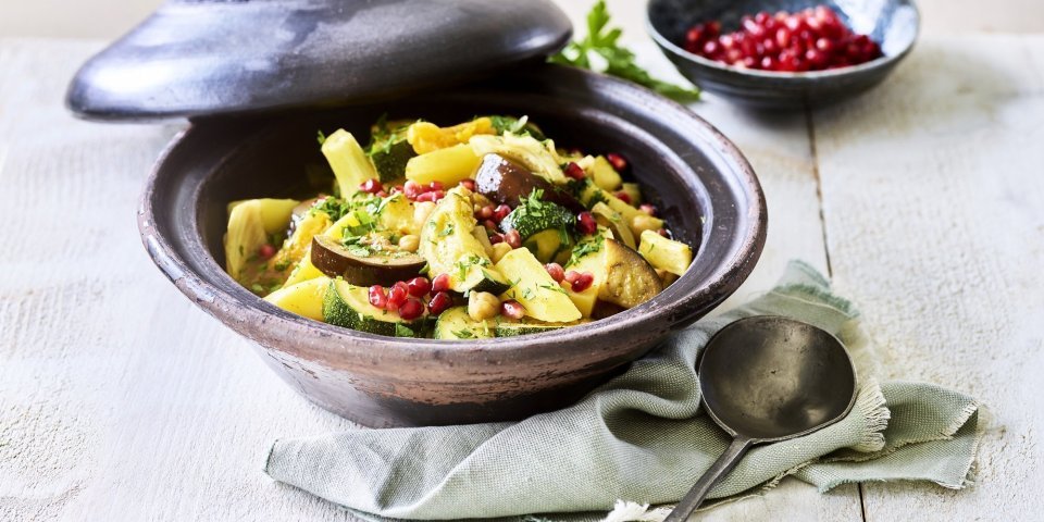 Er staat een tajine op tafel, de tajine heeft een zwarte kleur en is gevuld met stukjes aardappel, courgette, venkel, abrikozen, kikkererwten en granaatappelpitten. 