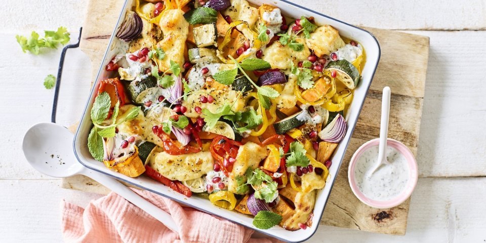 Op een witte tafel staat een houten snijplank met hierop een vierkante ovenschotel. Hij is gevuld met zoete aardappel, courgette, paprika en rode ui. Er is ook Berloumi gebruikt in dit gerechtje. 