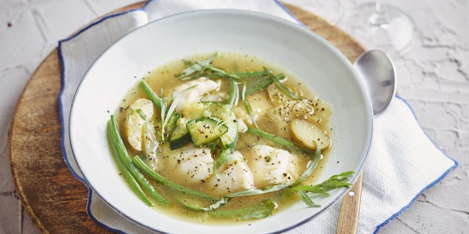 Een heerlijke maaltijdsoep is geserveerd in een wit soepbord. Deze staat op tafel, op een houte ronde snijplank, op een witte servette met blauwe rand. Het bord is gevuld met de doorschijnende bouillon met hierin de stukjes groenten en vis. Echt een feestsoepje. 