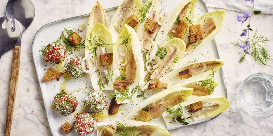 Op een rechthoekige dienschotel liggen meerdere blaadjes van witloof gevuld met een stukje sprot, ringen van sjalot en croutons. In het hoekje van de schotel liggen de roomkaasballetjes, deze balletjes zijn gerold in roze peperkorrels en verse dille waardoor ze als het ware een jasje van kruiden kregen.