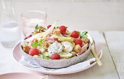 Een rijkelijke aardappelsalade is geserveerd in een grote kom. Je ziet de aardappel met de dressing, stukjes Breydelham, framboosjes en appel. De kom staat op een pastelkleurig roos bord. Er staat ook een karaf water langs met een glas in dezelfde tint als het roze bord. 