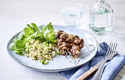 Op een lichtblauw groot bord liggen 3 gegrilde brochettes, we serveren ze met een couscoussalade van erwtjes en verse kruiden. Naast het bord ligt een blauwe servette met bestek langs. Er staat ook een glaasje water bovenaan het bord met een flesje water naast.  