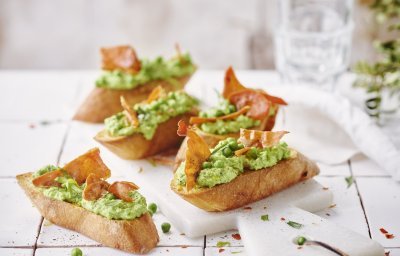 Vijf klaargemaakte bruschetta liggen op tafel. De spread van geitenkaas en erwten kleurt mooi groen, de bruchetta werd afgewerkt met een stukje krokant gebakken gedroogde ham. 