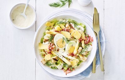 Een rijkgevulde salade staat klaar op tafel. Het witte bord is gevuld met sla, gekookte eitjes, stukjes asperges en kaasschilfers. Dat is de lente proeven in je bord, als je het mij vraagt!