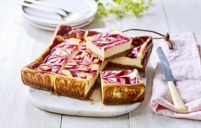 Een gebakken kaastaart staat op een marmere snijplank, het is wel een speciale cheesecake. Nl, de bovenzijde bevat frambozencoulis, waardoor er bovenaan een marmerpatroon is van de kaas en framboosjes. Echt een pareltje om naar te kijken. De taart is al aangesneden om onmiddelijk aan te vallen. 