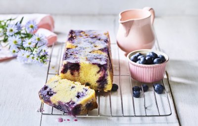 Op een bakrooster ligt de citroencake af te koelen, hij heeft een prachtige kleur door de blauwe bessen die tinten van paars achterlieten in de smeuige cake. Er staat nog een extra portie blauwe bessen naast voor extra fruitplezier.