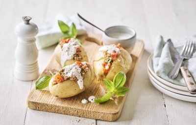 Enkele klaargemaakte en afgewerkte gepofte aardappelen liggen op een houten snijplank. Ze zijn gevuld met gehakt, stukjes tomaat en afgewerkt met een geitenkaasdip. Naast de aardappelen staan enkele bordjes, bestek en servetten om meteen te beginnen. 