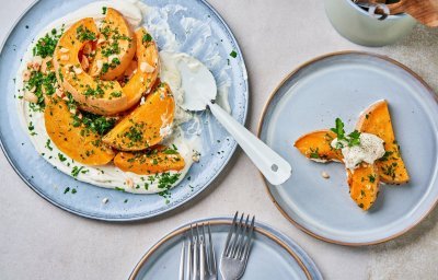 Er staat een lichtblauw-grijs bord waarop een kaasspread is uitgesmeerd. Hierop liggen stukken geroosterde butternut. Het recept is afgewerkt met fijngehakte peterselie en noten.