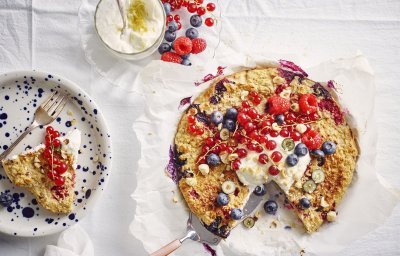 Een aangesneden taart staat op tafel met een wit laken er op. De kleuren van het rood fruit geven hierdoor nog een mooier zomers kleur. Naast de havermouttaart staat een bordje met blauwe spikkels en hier het stukje taart erop en een glaasje met de topping van de platte kaas en citroenzeste. 