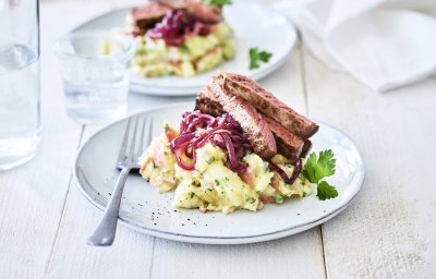 Een lichtblauw bordje is gevuld met een combinatie van puree en appelmoes. Deze puree is afgewerkt met gebakken rode ui en stukjes vlees. Er staan 2 van deze bordjes op tafel met een glas water. 