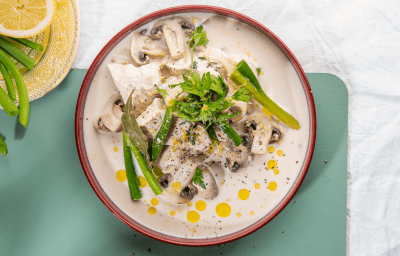 Kip gegaard in melk staat op tafel in een diep beige bord met bordeau randje. Er liggen lente-uitjes en champignons bij. Het bordje staat op een blauwgroene onderlegger. 