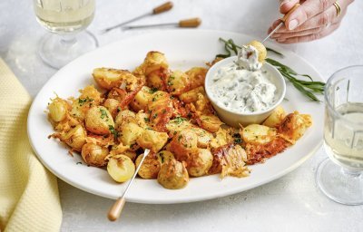 Een groot wit bord staat midden op de tafel, het is gevuld met heerlijke krokante aardappeltjes met kaas en een dipsausje van dragonmayonaise. Er is een hand die er eentje prikt en er staan twee glaasjes wijn. De aperitief is gestart. 