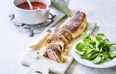 Een rol bladerdeeg ligt op een marmere snijplank. Het bladerdeeg is al aangesneden en zo zie je de spinazie en het vlees zitten. Wij serveren hem in kleine stukjes, net zoals een sushi, met homemade tomatensaus. 