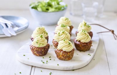 Gehaktbroodjes die klaargemaakt zijn in een muffinvorm staan recht op een marmer snijplankje. Ze zijn afgewerkt met een toefje aardappelpuree dat er werd opgespoten. Ze zijn een feest om naar te kijken.