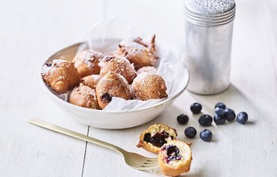 In een diep wit bord ligt een wit boterpapiertje met hierin enkele oliebollen. Er is eentje uitgeprikt en ligt op tafel doormidden gesneden, hier zie je duidelijk de blauwe bessen in zitten. Er is bloemsuiker over de oliebollen gestrooid voor een extra zoete toets. 