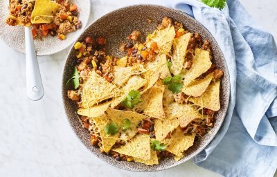 Onderaan de ronde ovenschaal ligt de chili con carne met bovenop de nachos. Er is al een portie uitgeschept op een wit bordje en staat langs de ovenschaal. Er ligt ook een blauwe servette en enkele blaadjes kroiander voor de kleur.