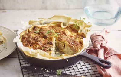 Op het aanrecht ligt een zwarte rooster met hierop de quiche in een gietijzeren pan. Hierbij ligt een roze servet, mes en bordje waarop al een stukje quiche op ligt. De quiche is afgewerkt met enkele verse blaadjes oregano. 