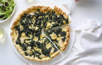 Op een taartschaal ligt de aangesneden quiche. Er is een witte taartschep onder één stukje quiche om naar een bordje te gaan. Langs de quiche staat ook nog een portie gemengde sla, voor nog een extra portie groenten. 